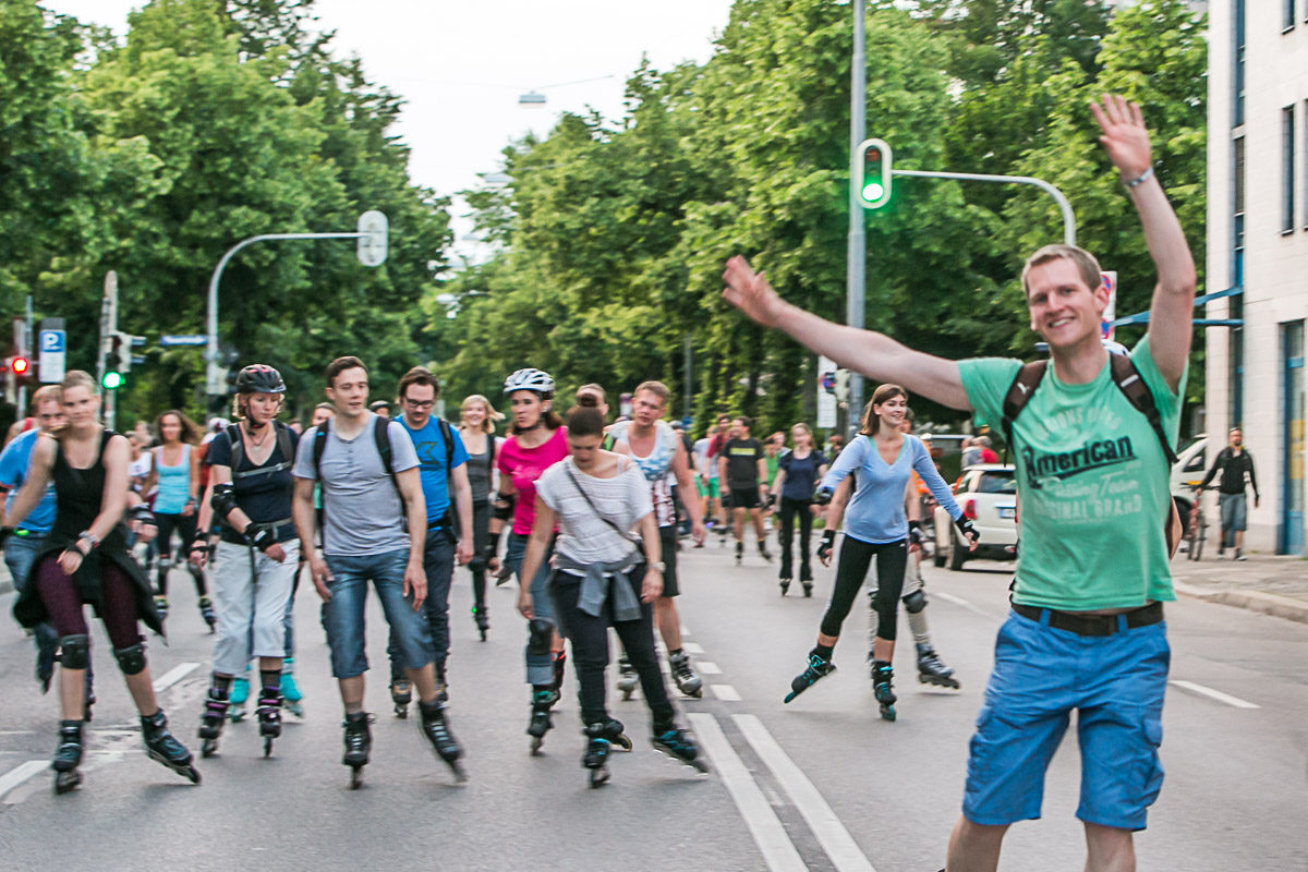 Group of people rollerblading down a city street; a man in a turquoise shirt in the foreground waves at the camera.