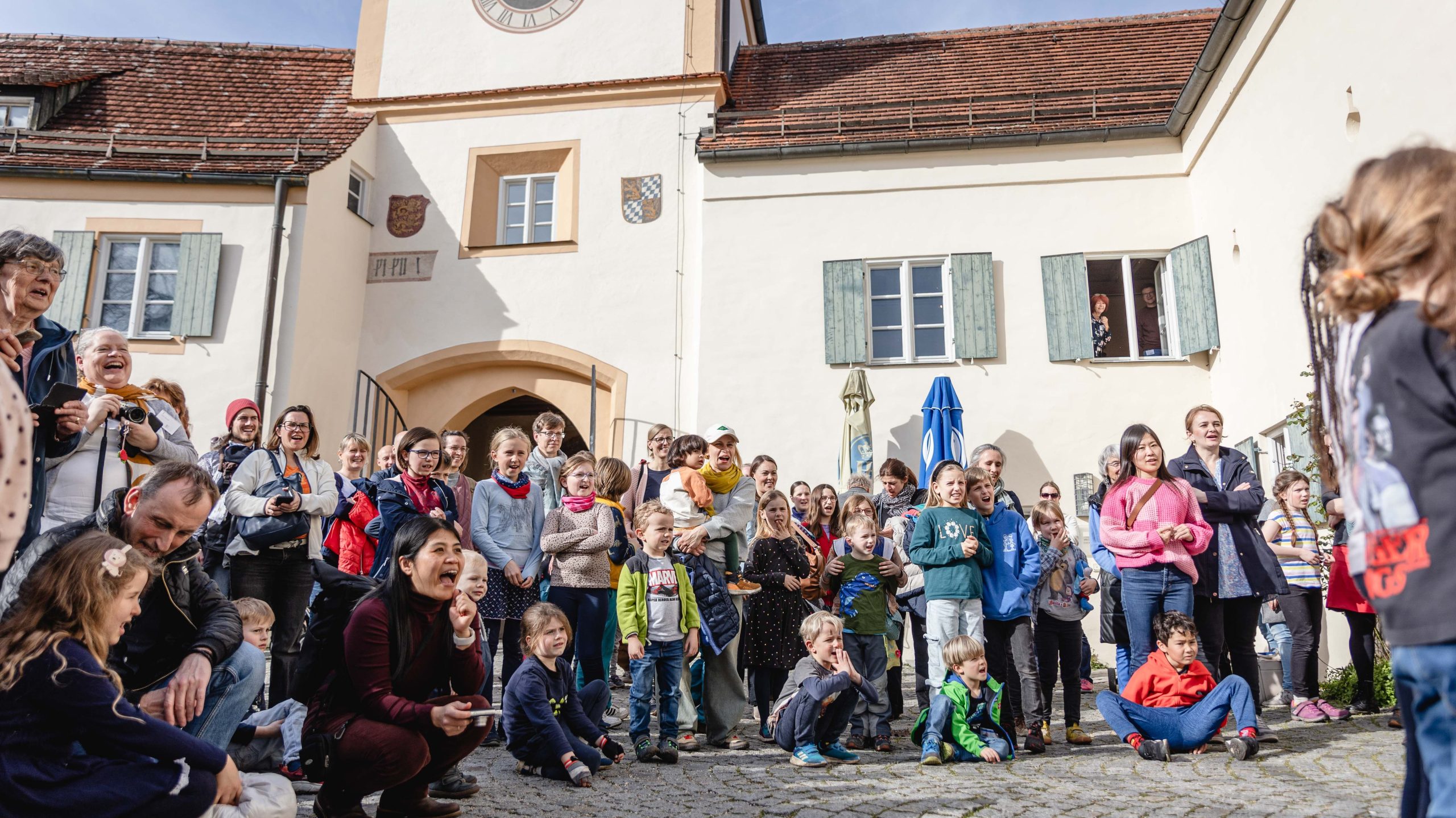 Crowd of children and adults cheering in a sunny town square, with a clock-tower building in the background.