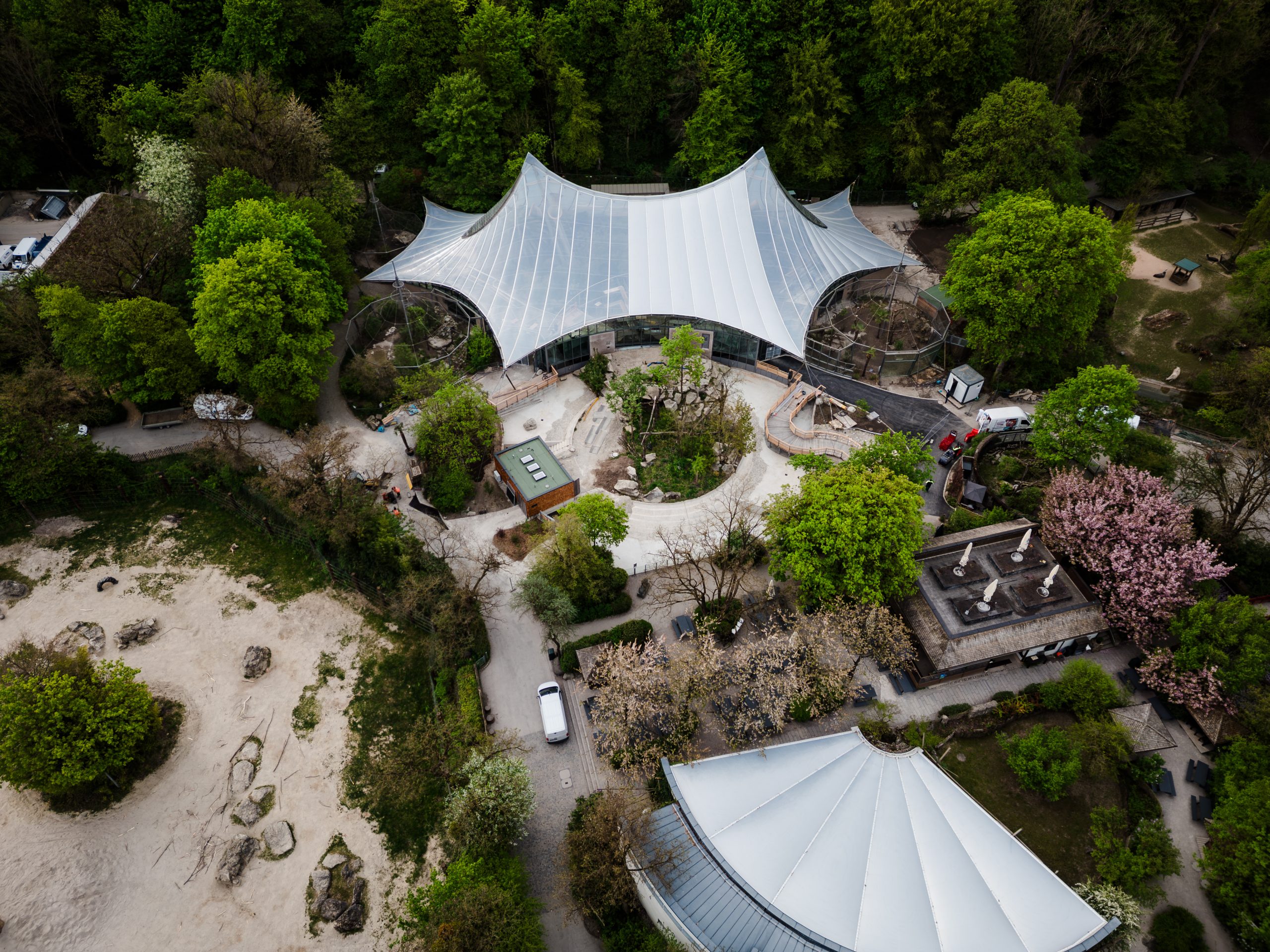 Aerial view of a park pavilion with a white sail-shaped canopy surrounded by trees and paths in a green area.