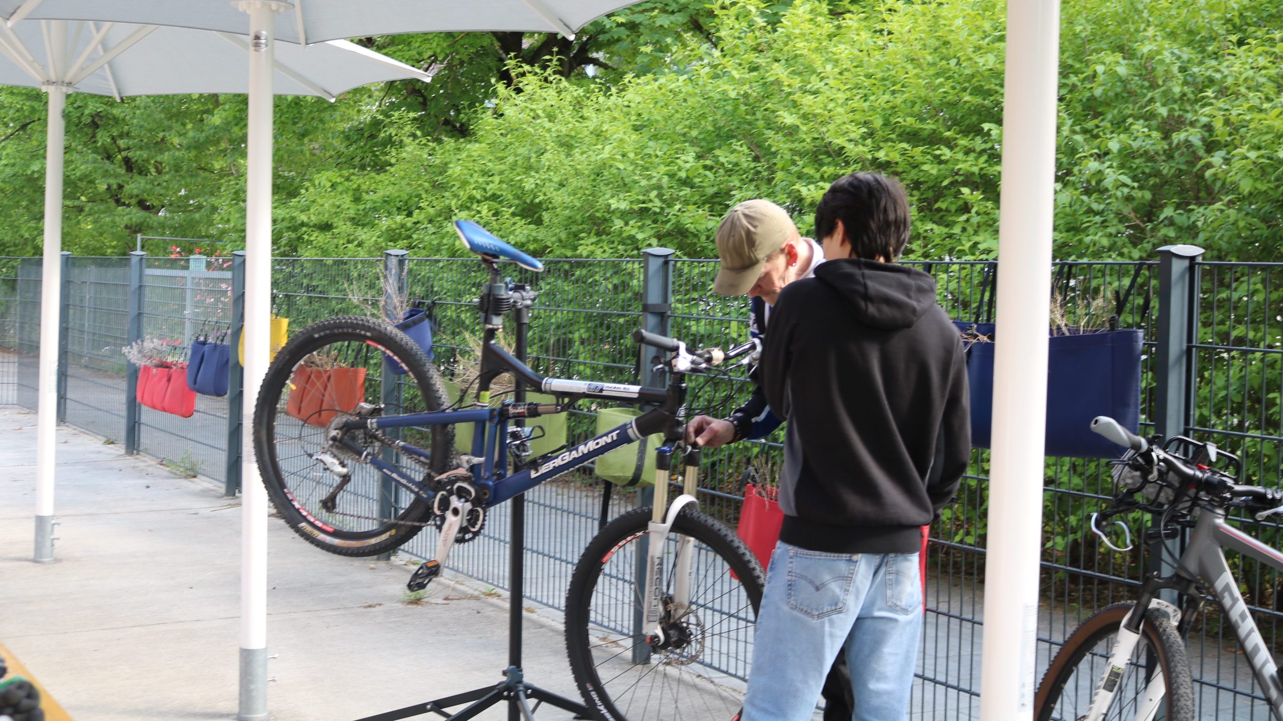Two people repair a blue mountain bike under white patio umbrellas, outdoors by a fence with green trees in the background.