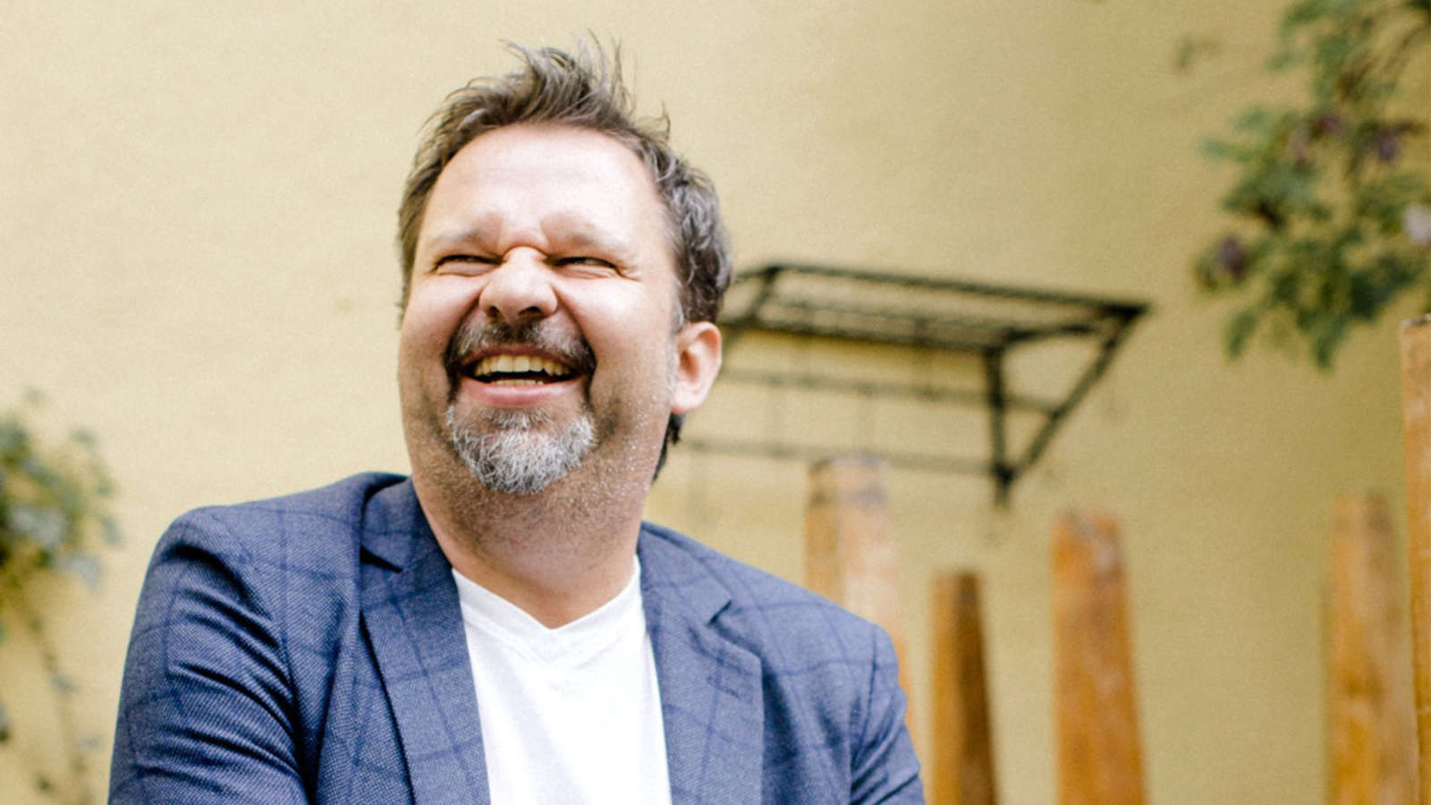 Man in a blue blazer laughing outdoors in front of a beige wall.