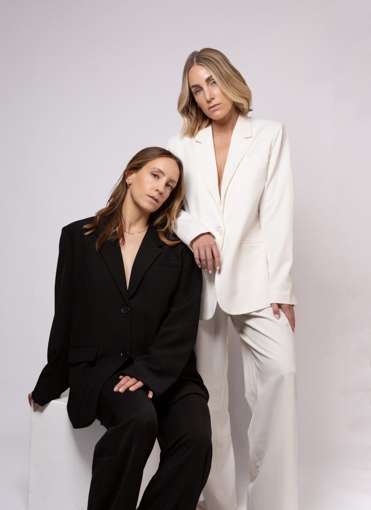 Two women in tailored suits pose together: one seated in a black blazer, the other standing in a white suit against a pale backdrop.