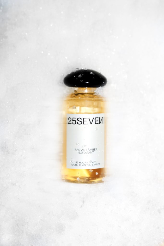 Small amber glass bottle with a black cap, partly buried in snow; label reads 25SEVEN and Radiant Amber Exfoliant.