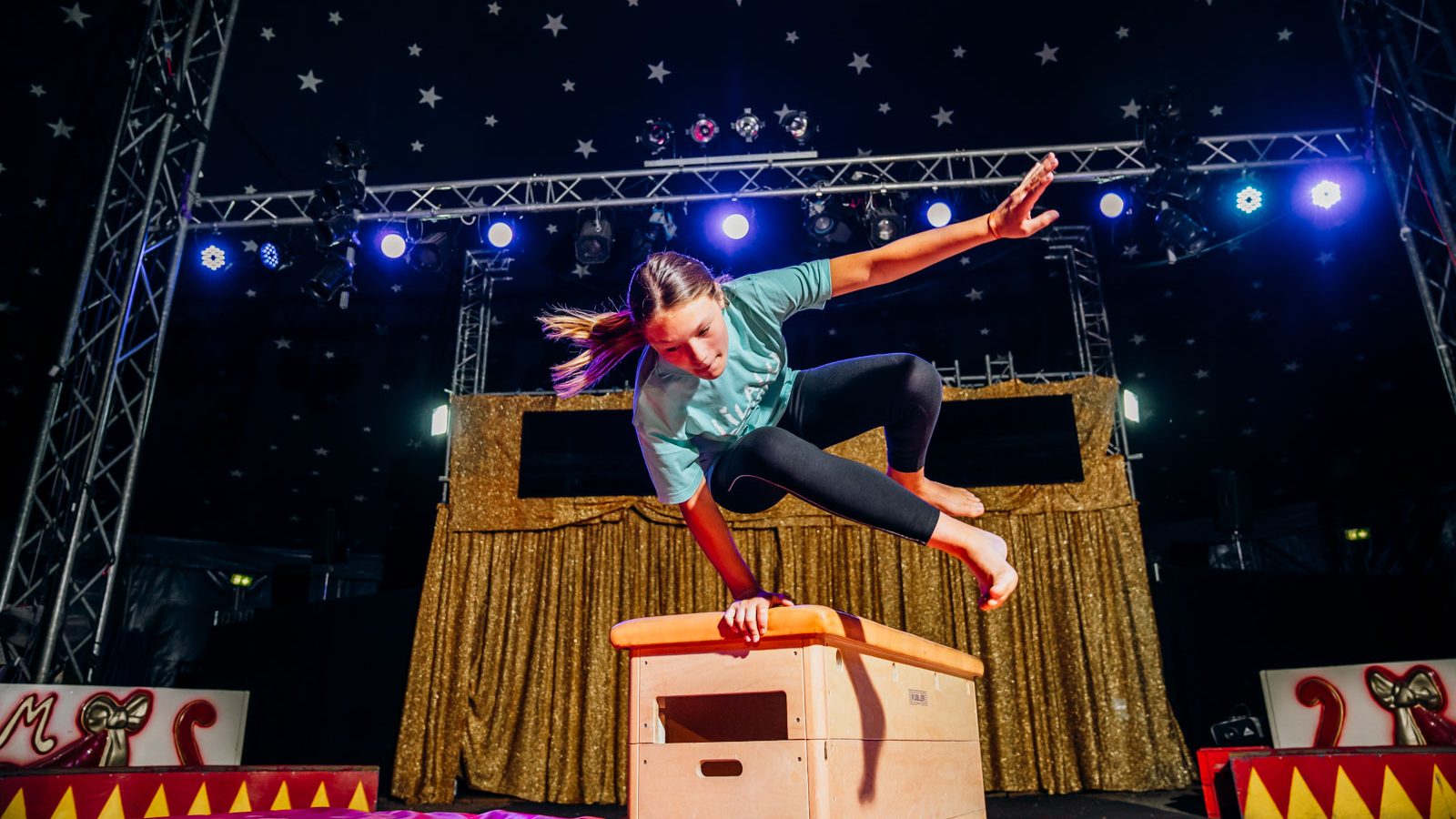Female gymnast mid-air while vaulting over a wooden box on a carnival-style stage with gold fringe backdrop and starry ceiling, blue stage lights