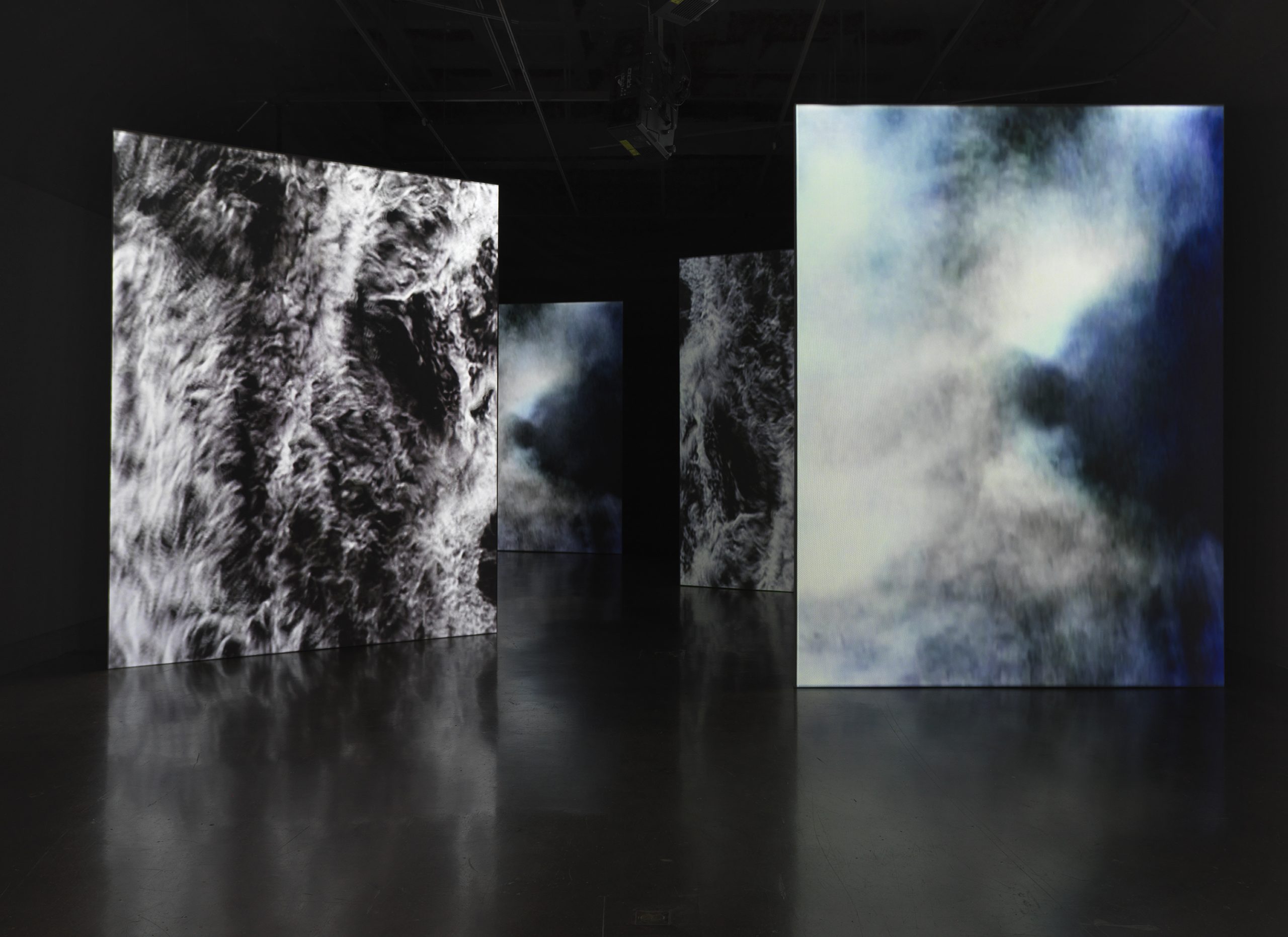 Tall vertical screens in a dark gallery displaying abstract smoky textures in black-and-white and blue hues, part of an immersive art installation.