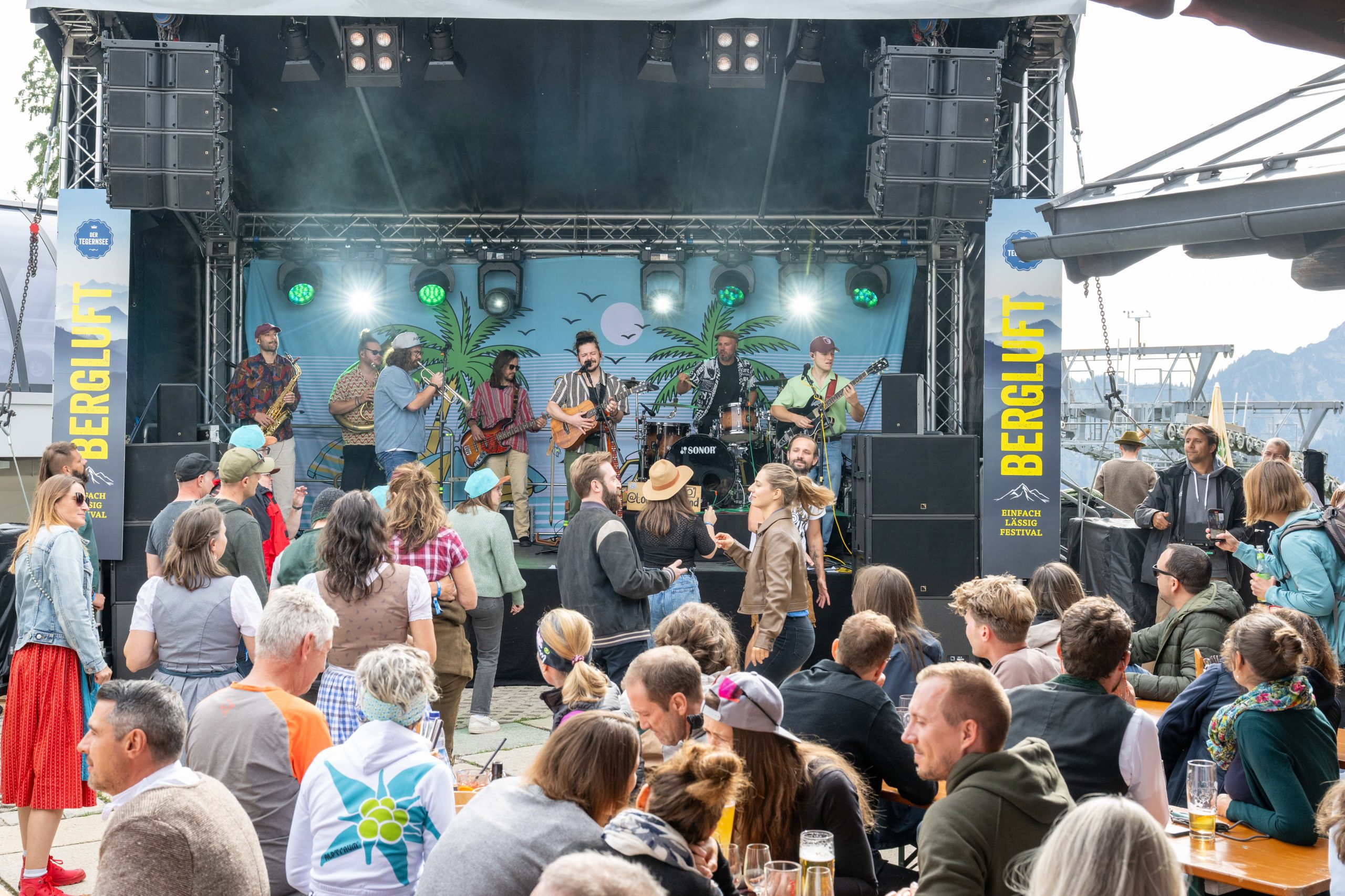 Outdoor music festival stage with a live band playing and a cheering crowd in front of palm-tree backdrop and banners on the sides.