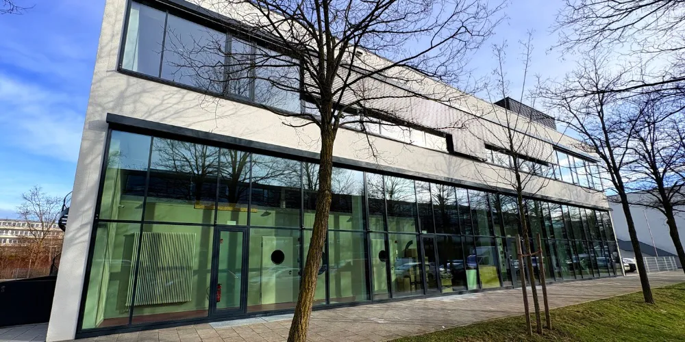 Modern glass-front office building with a paved walkway and leafless trees along the sidewalk.