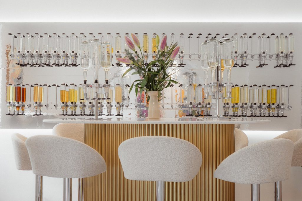 Atelier-Inspire-parfum - IN München - Das Stadtmagazin Row of glass test tubes filled with colored liquids on a laboratory-style shelf behind a chic bar, with a vase of pink lilies at the center and cream upholstered stools in the foreground.