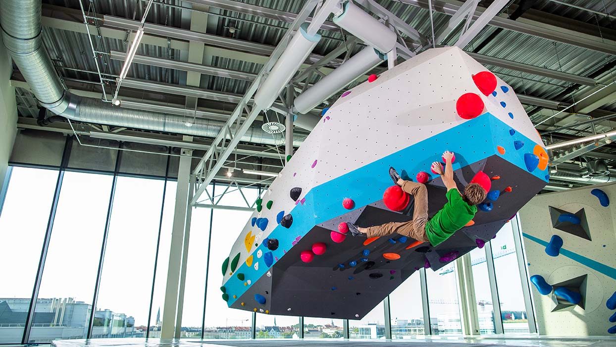 BW_Plaza_KursErwachsene-245 - IN München - Das Stadtmagazin Climber in a green shirt scales a large suspended white-and-blue climbing block with colorful holds inside a modern indoor gym.