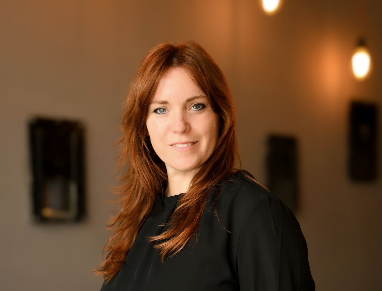 Portrait of a smiling red-haired woman wearing a black top, in a warmly lit indoor setting