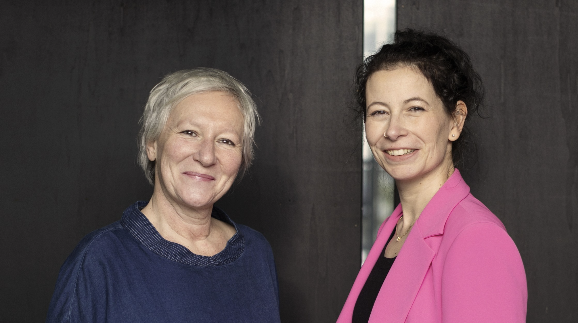 Two women standing side by side, smiling at the camera against a dark backdrop; woman on left wears a blue top, woman on right wears a pink blazer with a black top underneath.