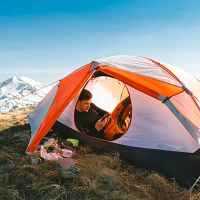 Camping - IN München - Das Stadtmagazin Person inside an orange-and-white tent on a grassy hillside, cooking with camping gear, with snow-capped mountains in the background.