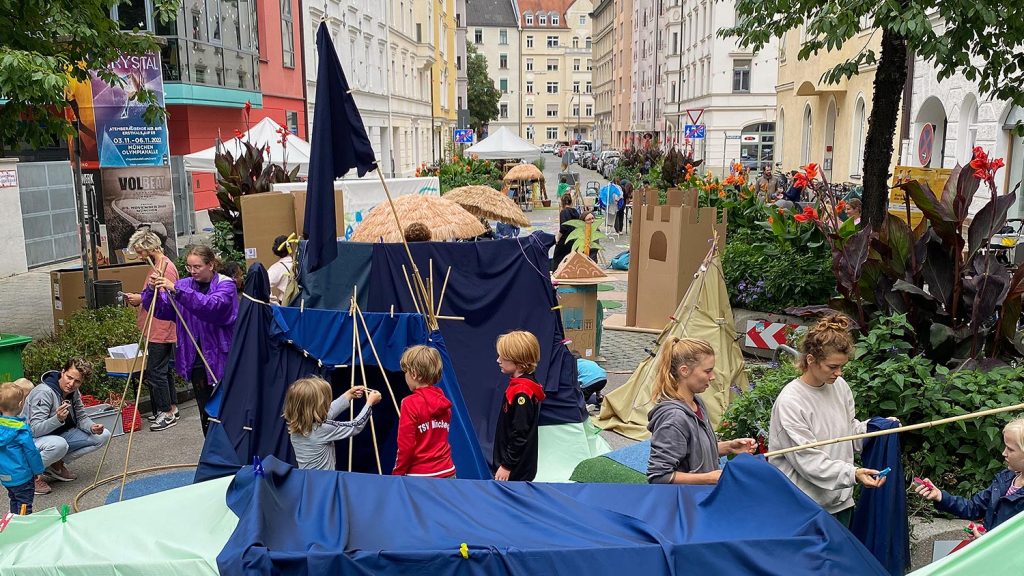 Street festival with children building a blue fabric tent and cardboard structures along a cobblestone street lined with colorful buildings.