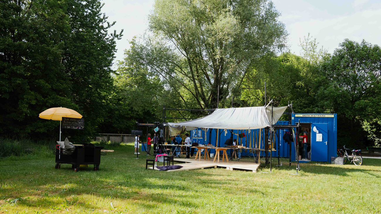 Outdoor event setup under a white canopy with wooden stools; blue portable containers in the background and people nearby in a grassy park area.