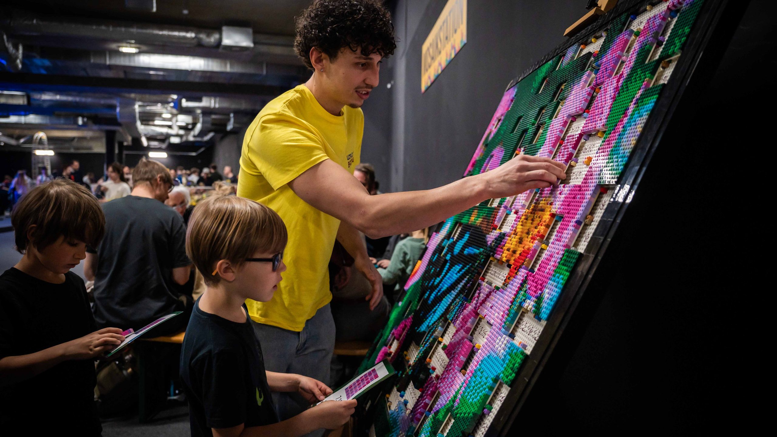 Man in a yellow shirt helps children place colorful beads on a large display board at a craft event.