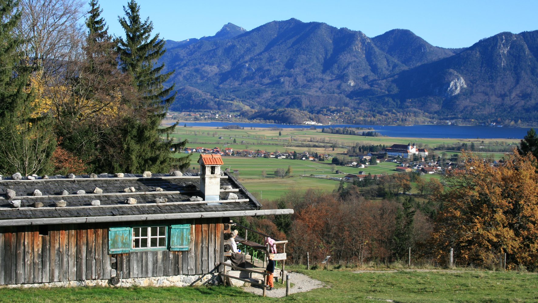 Rustic wooden house with green shutters in foreground, mountains and lake beyond, village in distance with people near deck.