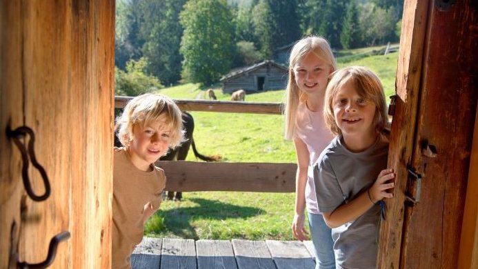 Three smiling children stand at a rustic wooden doorway, welcoming you to a sunny countryside yard with a fenced field beyond.