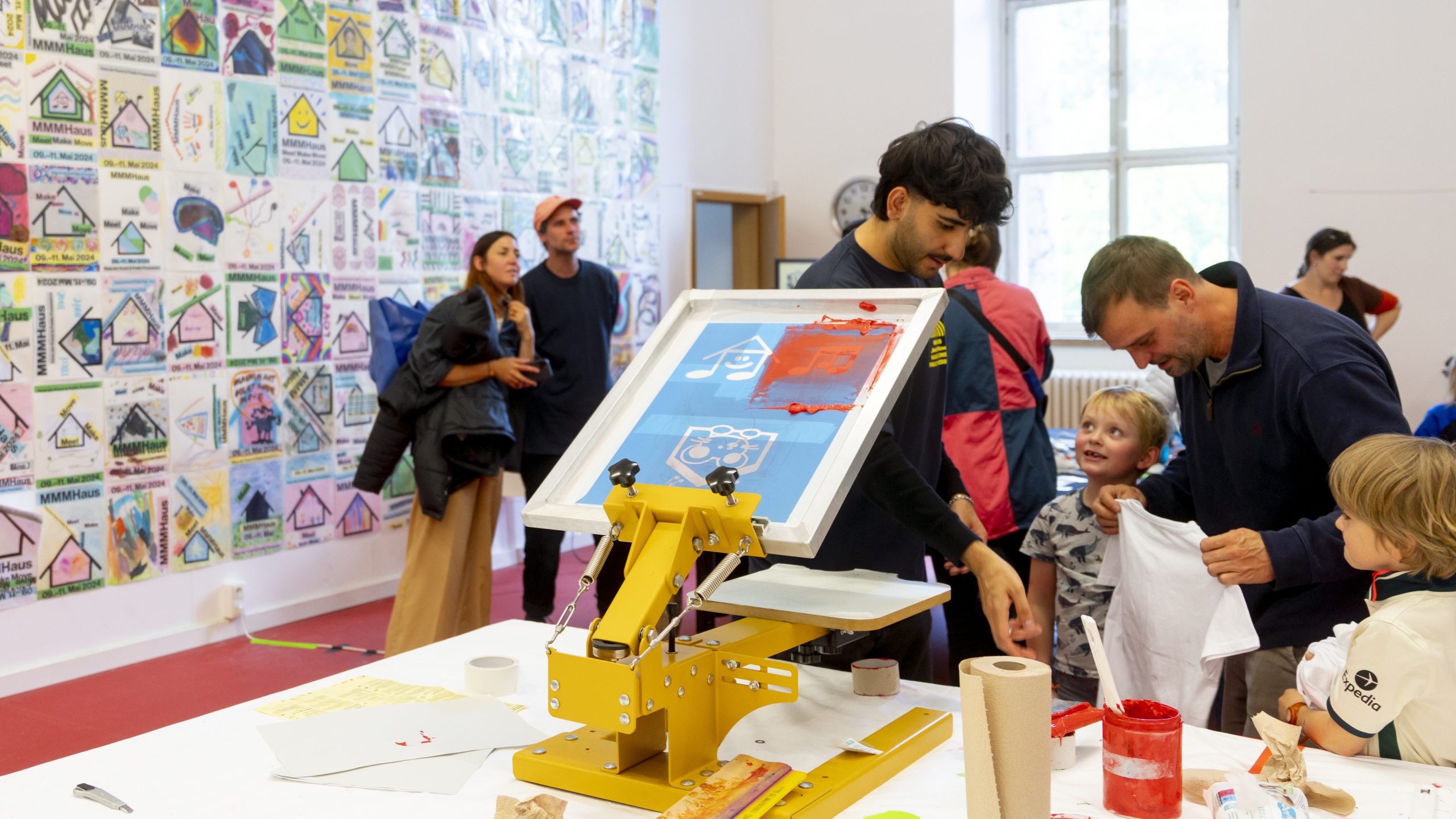People gathered around a bright yellow printing press at a workshop with a collage-covered wall in the background.