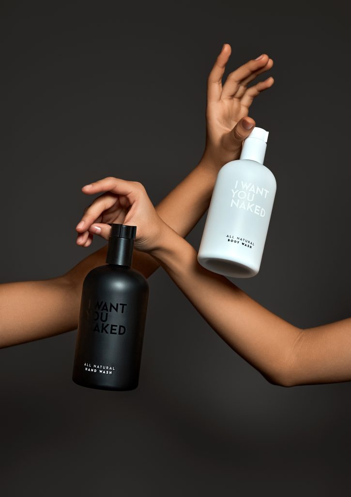 Hands hold and cross two pump bottles, a white bottle of 'I WANT YOU NAKED' body wash and a black bottle, against a dark backdrop. Text on bottles reads 'I WANT YOU NAKED' and 'ALL NATURAL BODY WASH'.