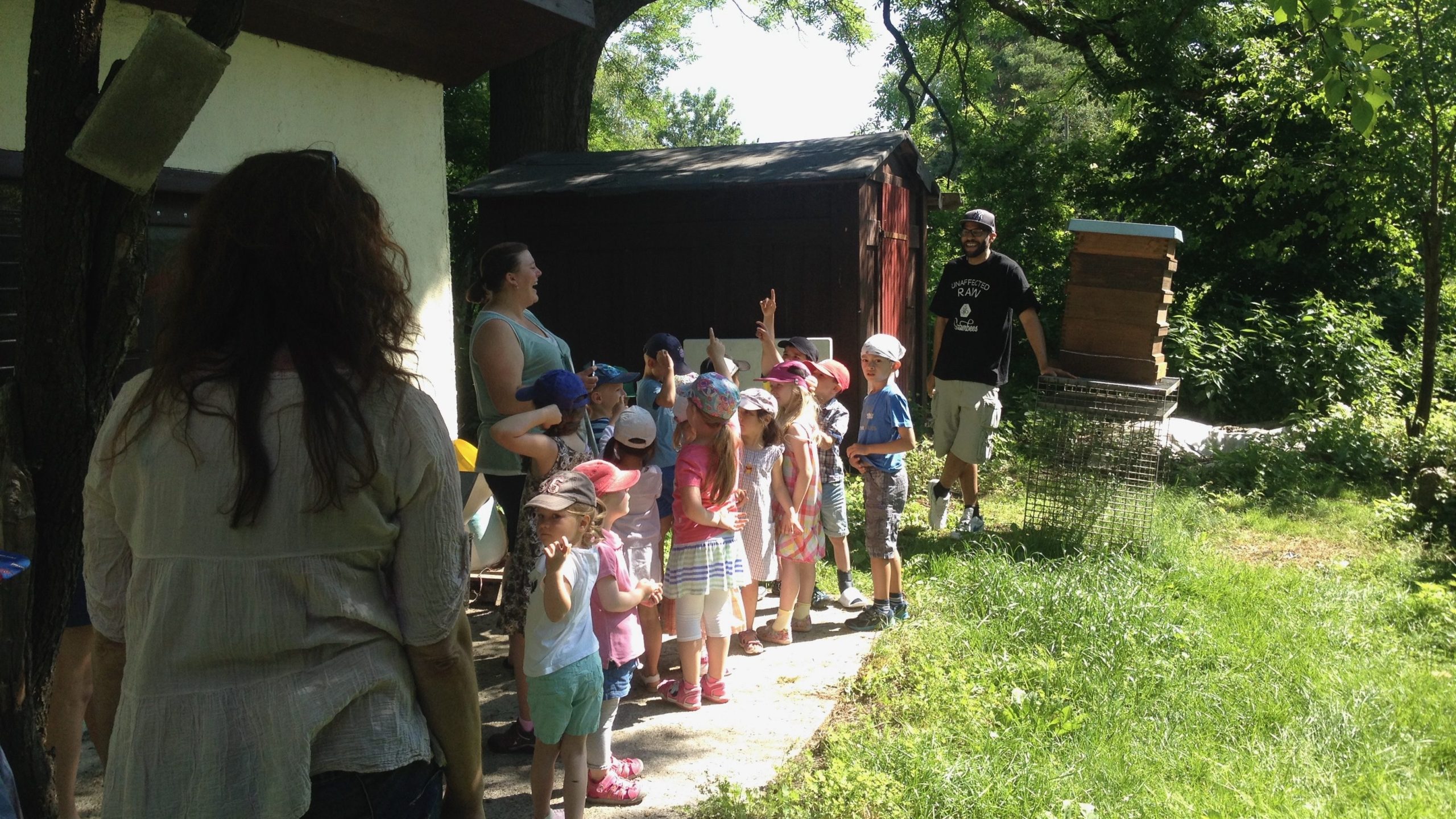 Children on a field trip listen to an instructor beside stacked beehive boxes outdoors on a sunny day.