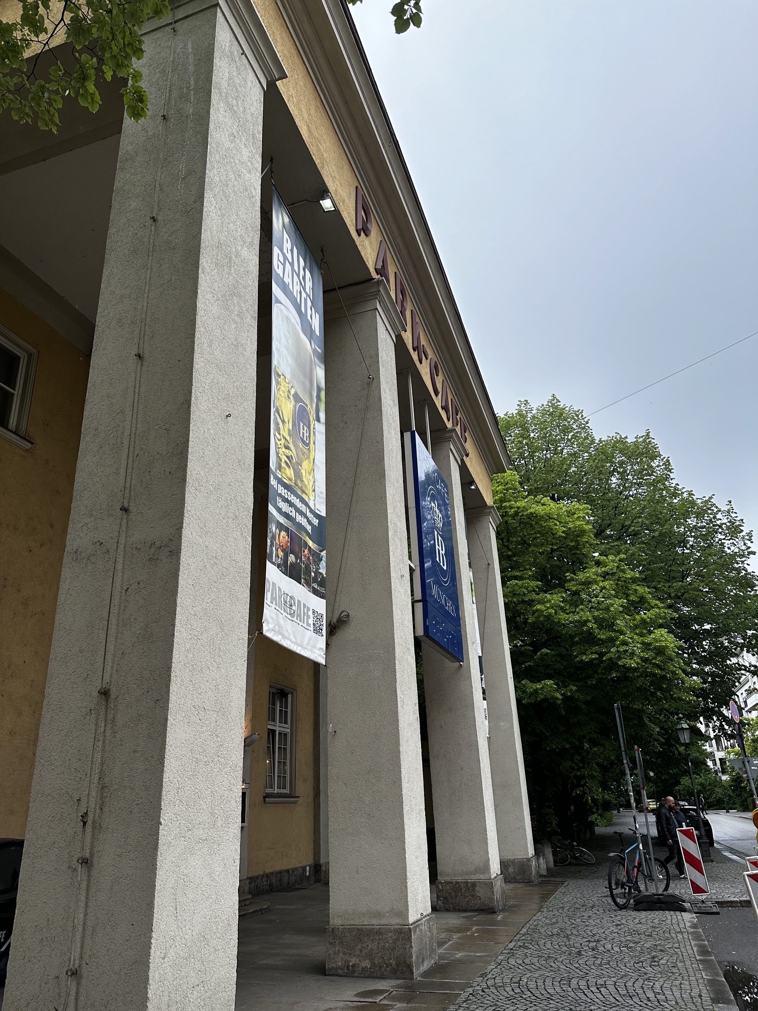 IMG_7157 - IN München - Das Stadtmagazin Facade of a building with tall concrete columns and hanging banners along a sidewalk area.