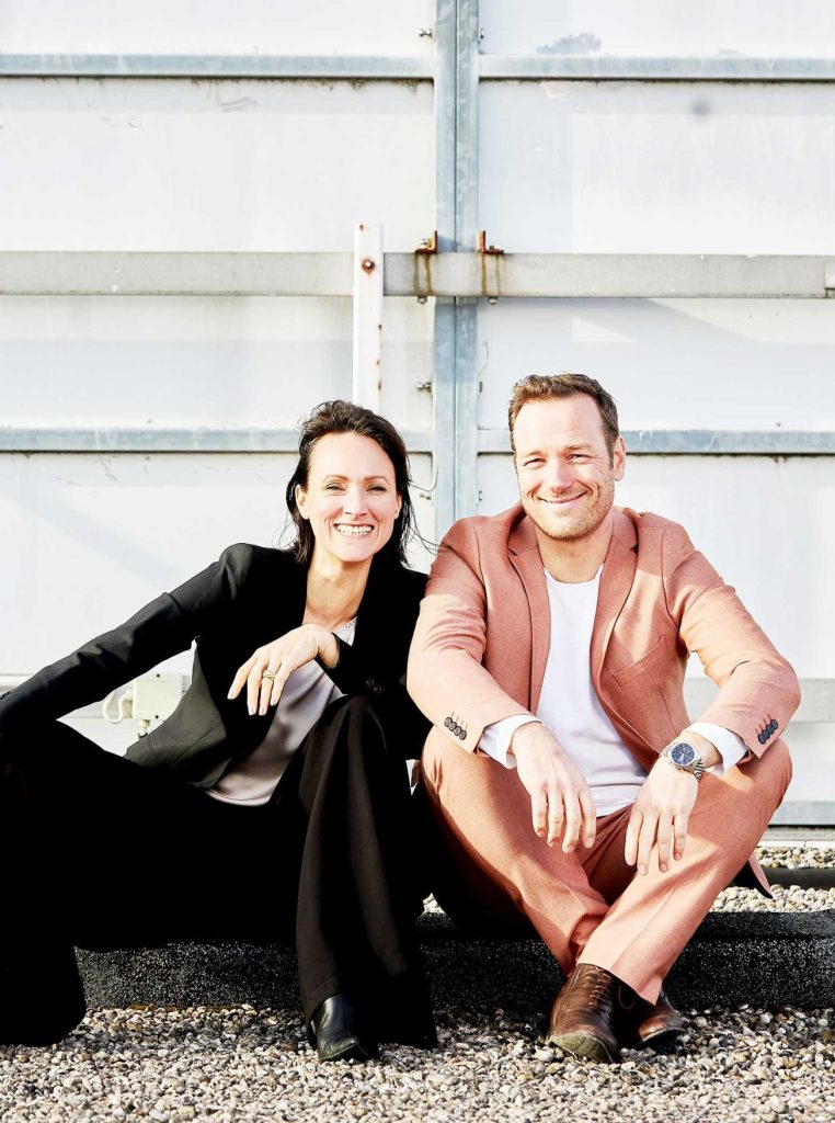 Two smiling adults sit on gravel in front of a large metal garage door, posing for a photo.”,