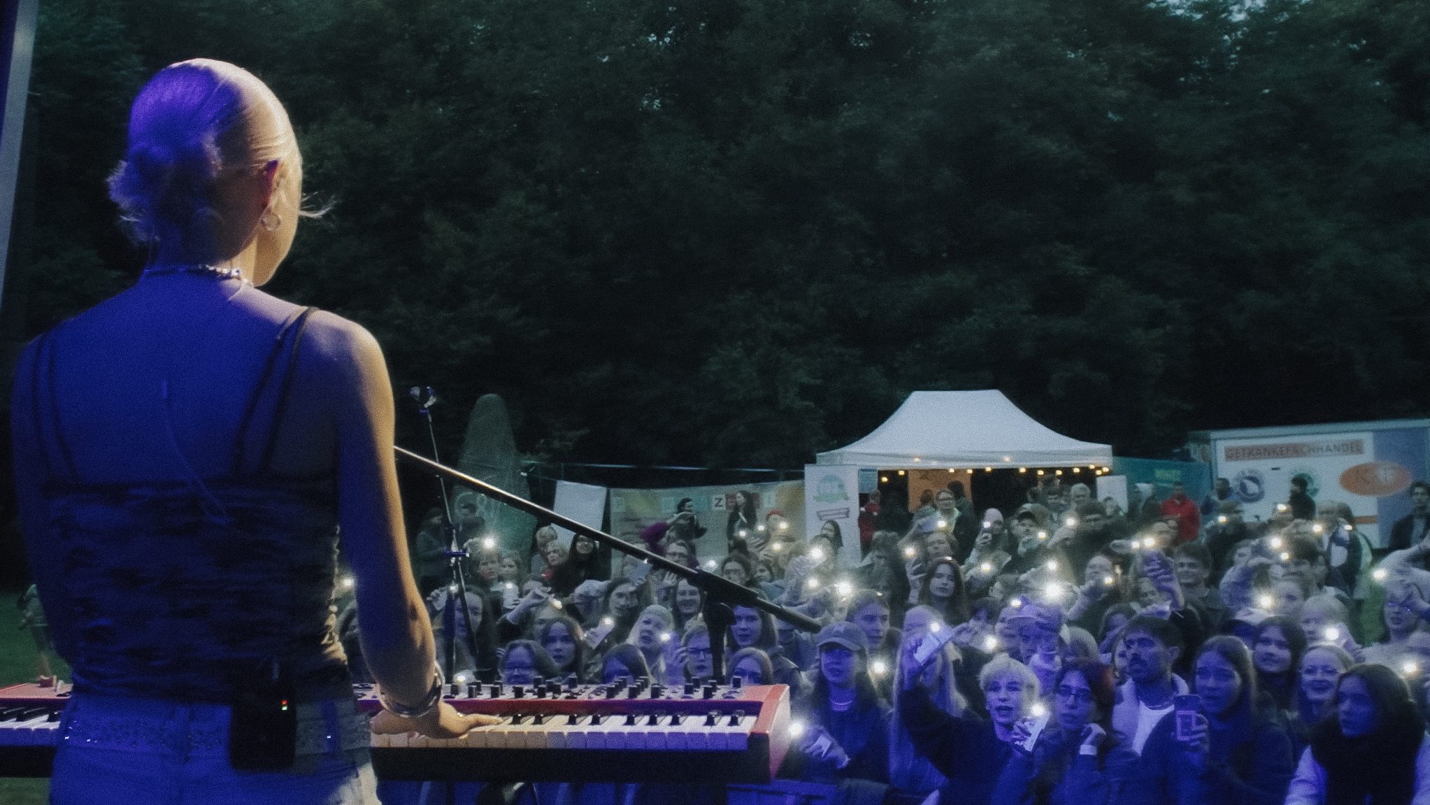 Back view of a musician in a blue outfit playing a keyboard on stage at an outdoor concert, a large crowd nearby with phone screens lit up.