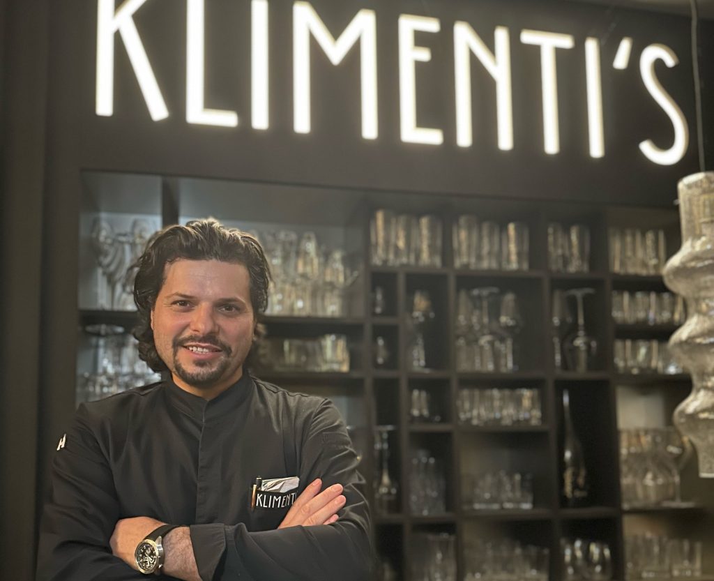 Klimentis - IN München - Das Stadtmagazin Man in a black uniform with arms crossed, standing in front of a glassware display in a store.