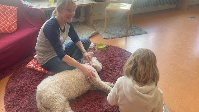 Smiling woman sits on a red rug stroking a fluffy white dog while a young girl watches in a bright living room.