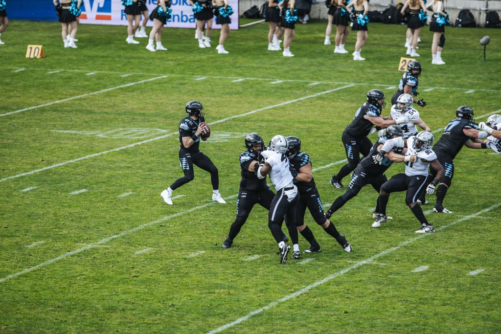 LewinWeinberger_2026_Week1_RAIatMUC_131 - IN München - Das Stadtmagazin Quarterback in black jersey holds football, backs to throw while teammates block defenders on a grassy football field behind him, cheerleaders in the background.