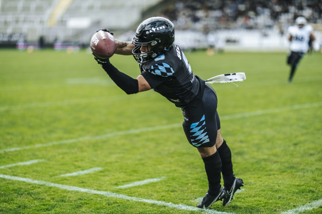 Rugby player in black-blue uniform lunges to catch a football on a green field, stadium blurred in background