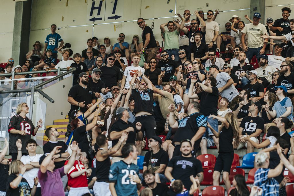 Crowd of fans in stadium stands cheering and raising arms during a game and crowding around.