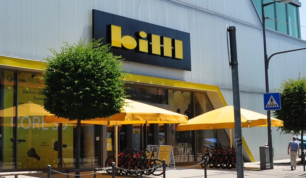 Front of a modern storefront with yellow awnings and umbrellas, bicycles parked outside, and a bold yellow-and-black sign above the entrance.