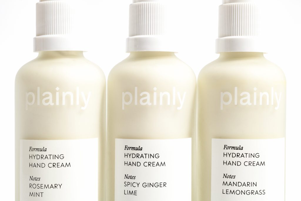 Three pale bottles of hydrating hand cream with white labels, showing scents like Rosemary Mint, Spicy Ginger Lime, and Mandarin Lemongrass.