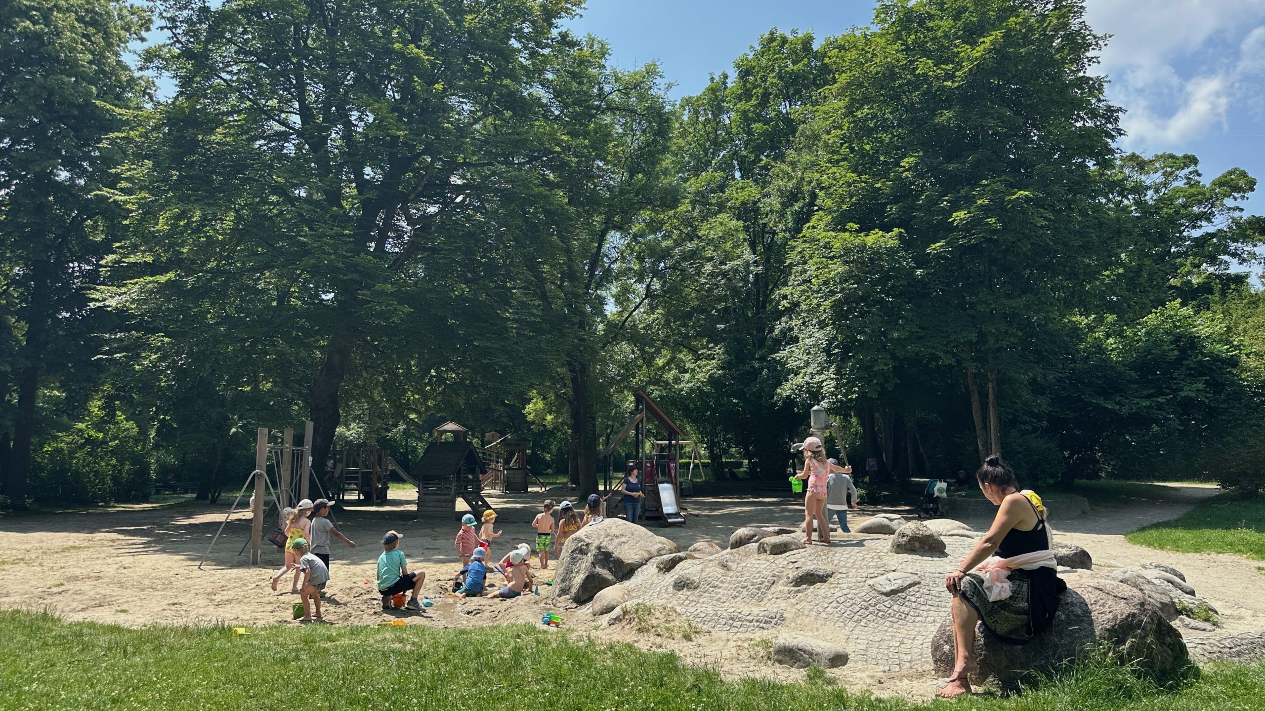 Children playing in a sandy playground under tall green trees, with adults supervising and large rocks nearby on a sunny day.