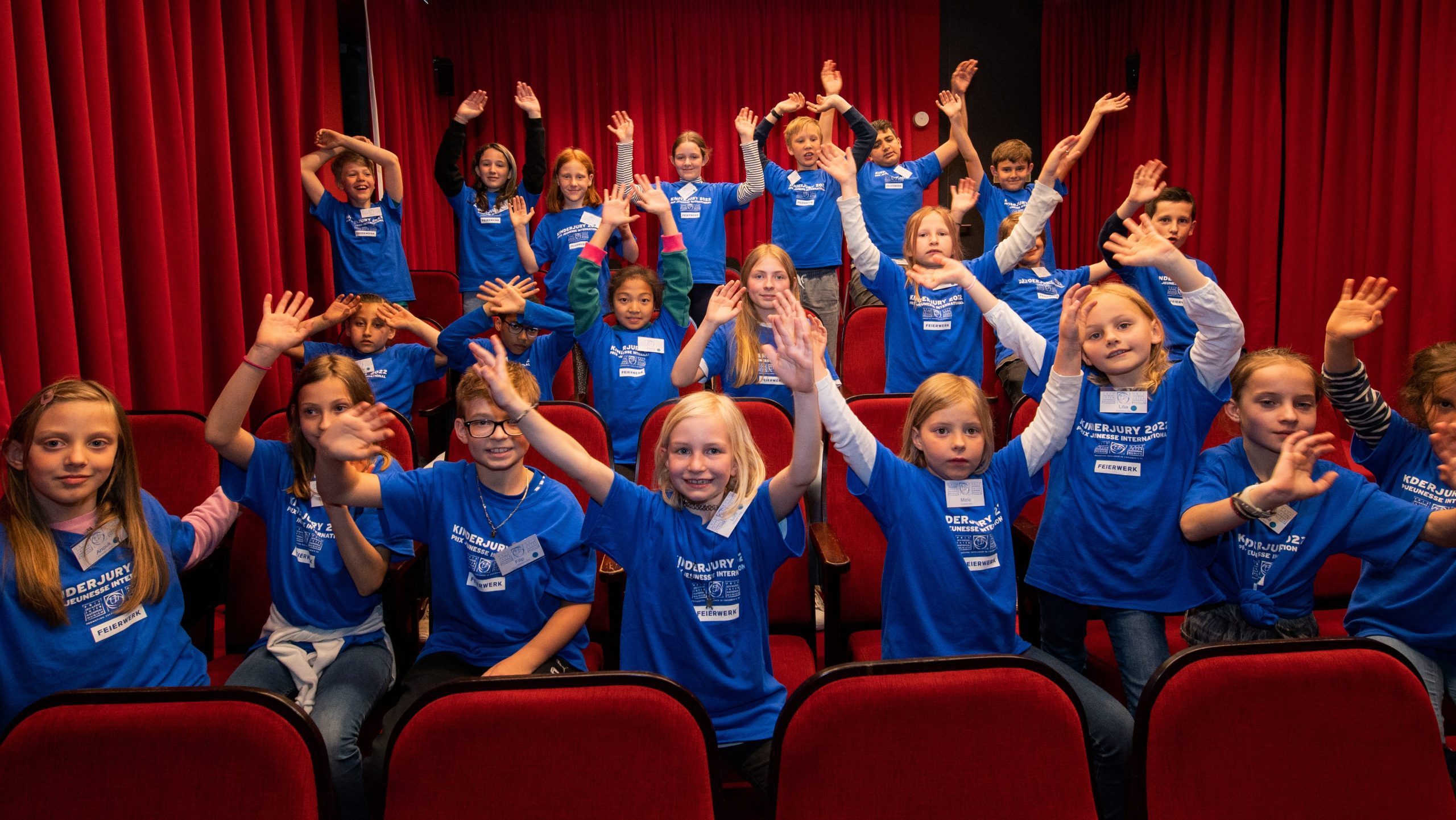 Group of children in blue shirts raising hands in a theater with red seats and curtains, posing for a group photo at an event.