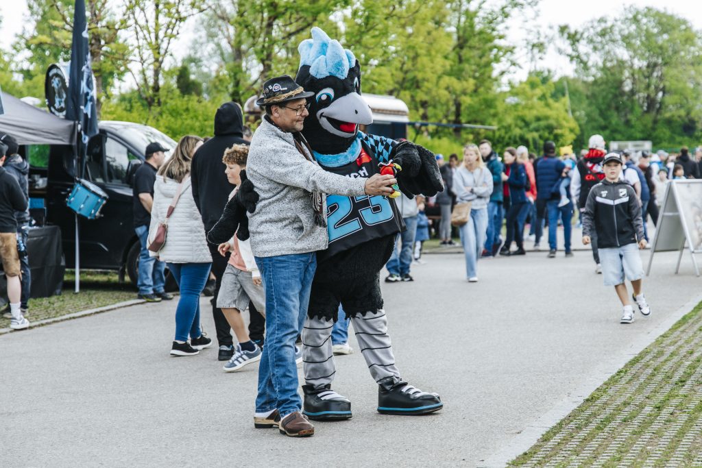Man dancing with a blue-crested mascot at an outdoor festival, crowd watching in the background.