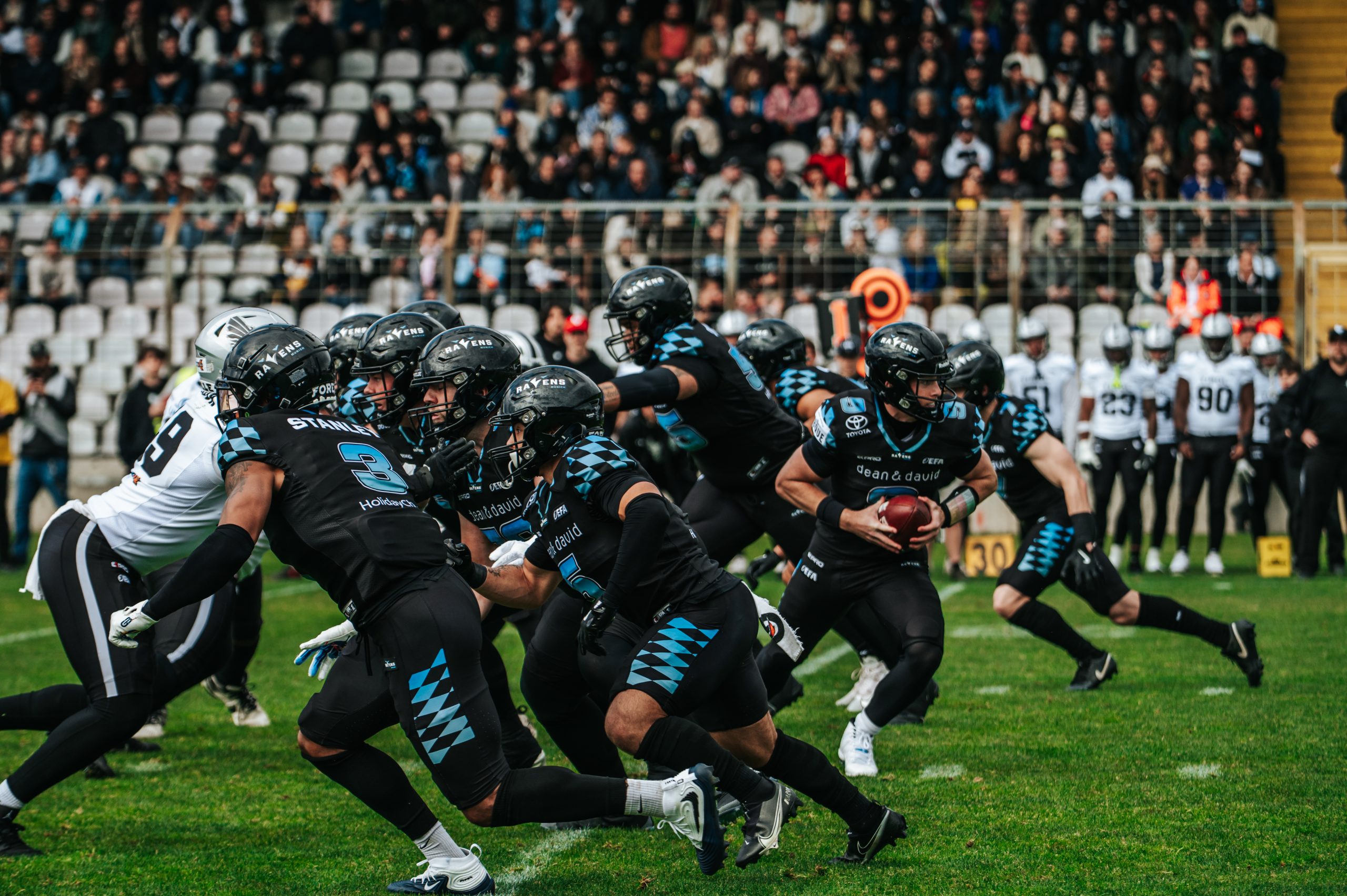 American football players in black and blue uniforms sprint the ball down the field as a crowd watches from the stands.