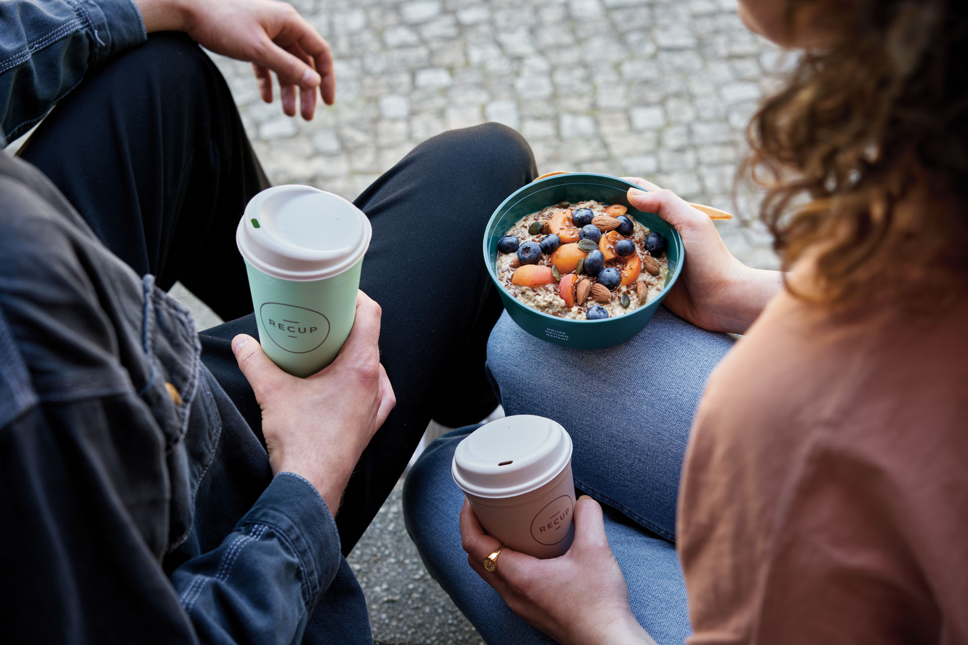 Two friends sharing a bowl of oatmeal topped with blueberries, apricots, almonds and seeds, while holding takeaway coffee cups outdoors.