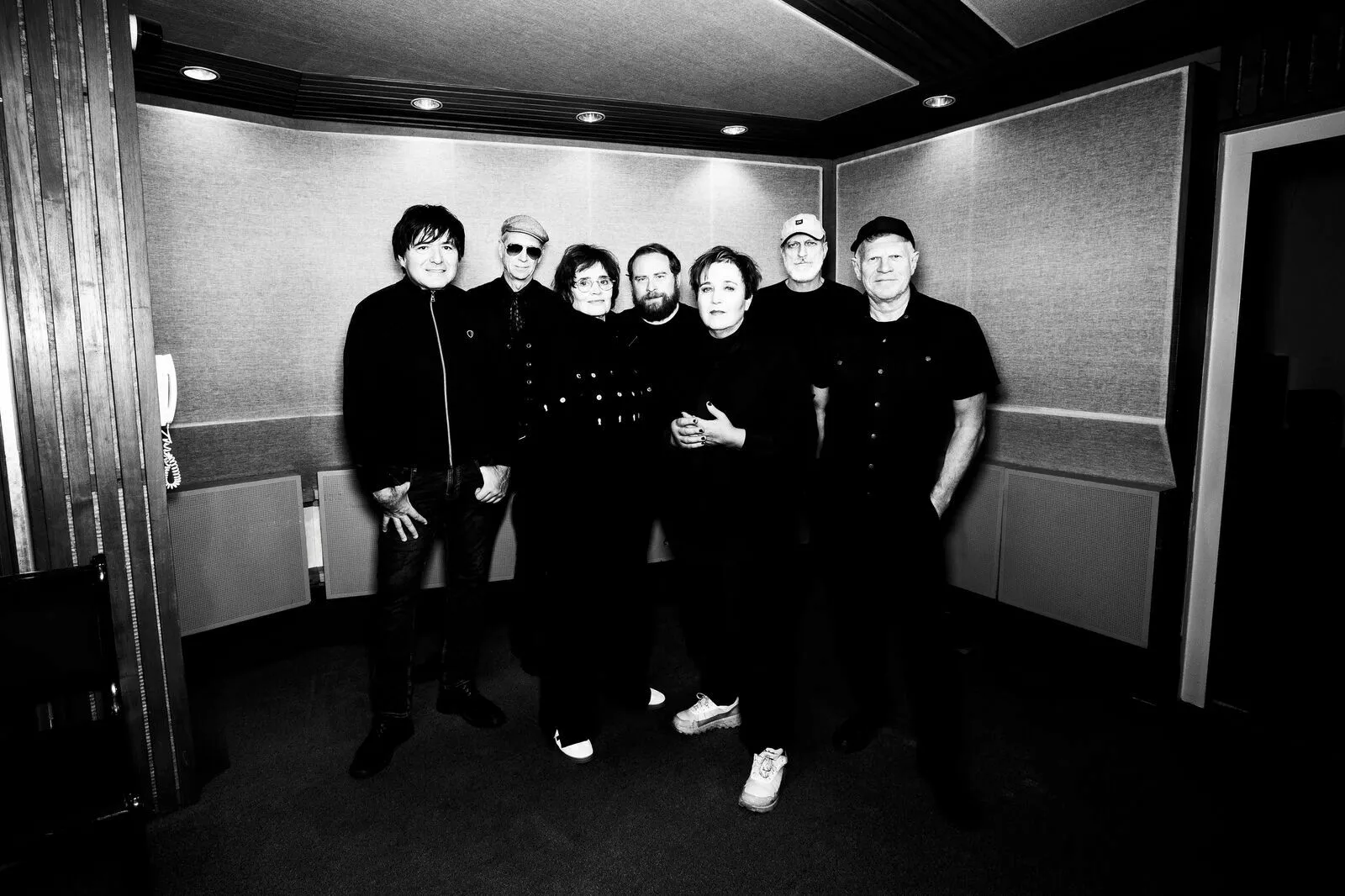 Eight people stand together in a recording studio, all dressed in black, posing for a group photo against padded walls and studio panels.