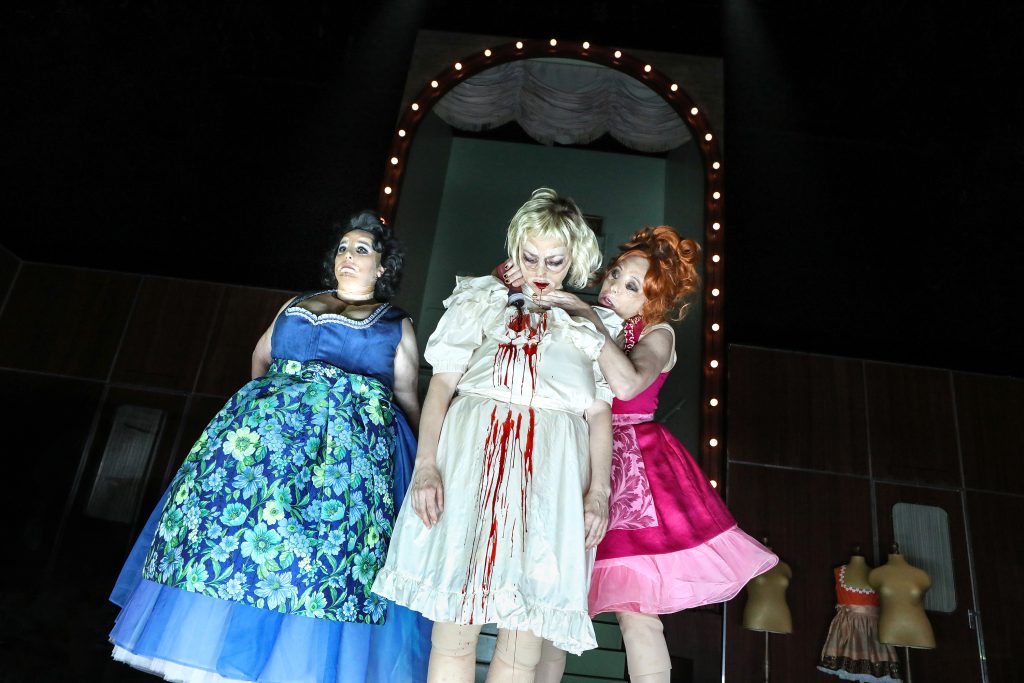 Die Präsidentinnen - IN München - Das Stadtmagazin Three drag performers on a stage wearing colorful gowns; central figure has blood-red accents on a white dress while others pose beside under a lit archway.