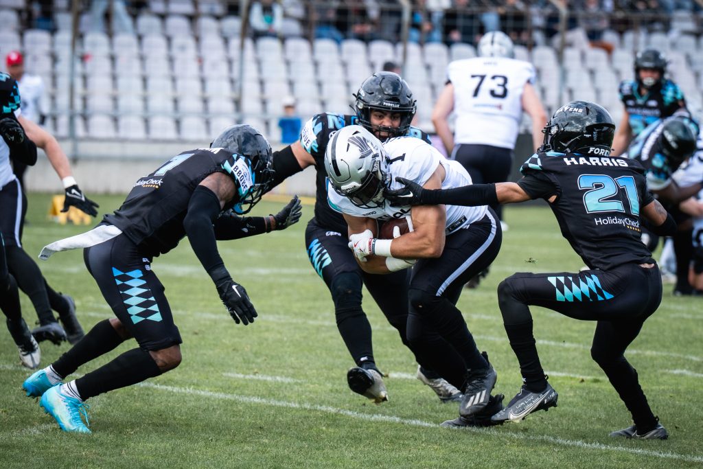 Players in black and teal uniforms tackle a ball carrier in white, forming a chaotic scrum on a grassy football field.