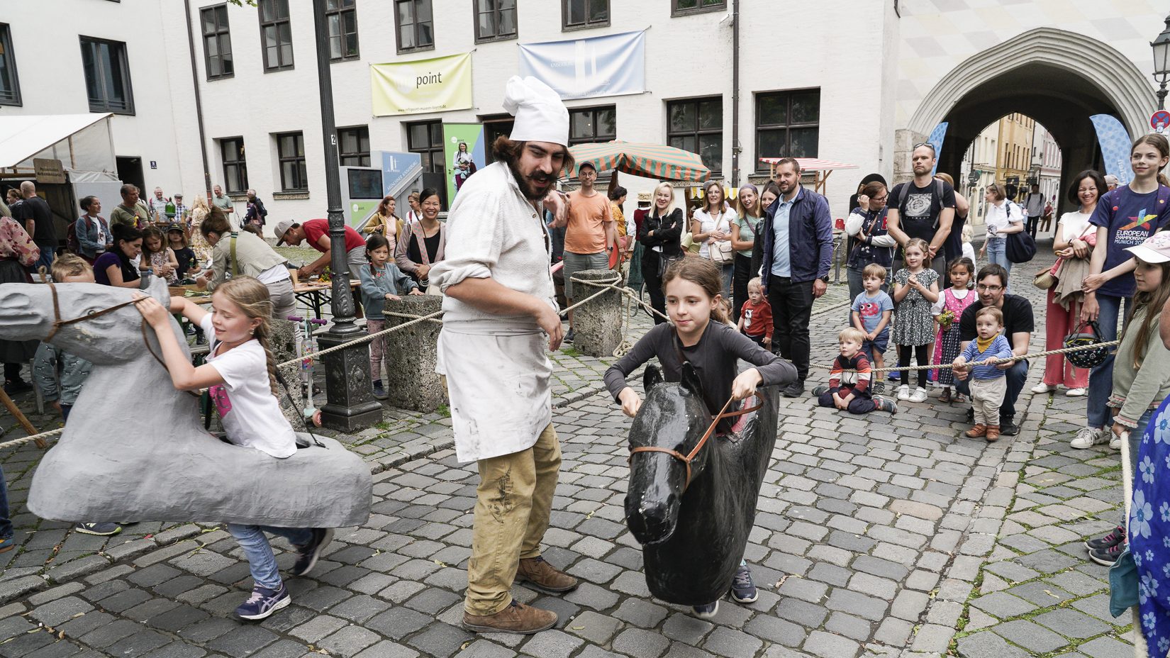 Children ride a black horse-shaped toy while a man in a white chef’s outfit guides them; a crowd watches on a cobblestone square.