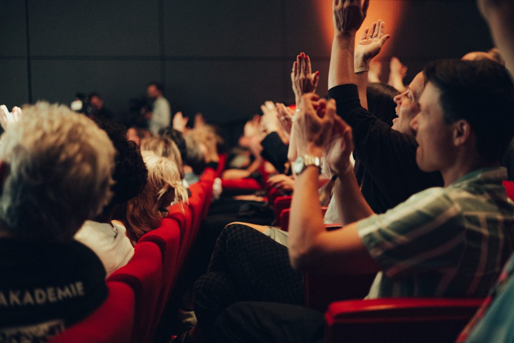 Crowd in a dim auditorium raises hands in excitement, red theater seats visible.