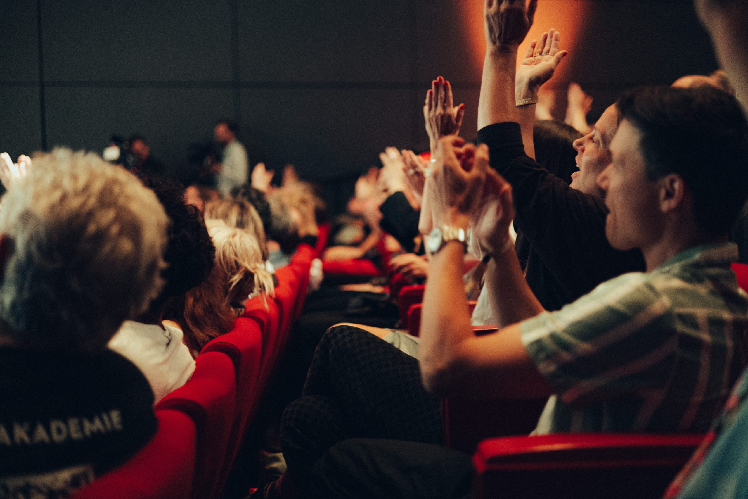 Crowd in a dim auditorium raises hands in excitement, red theater seats visible.