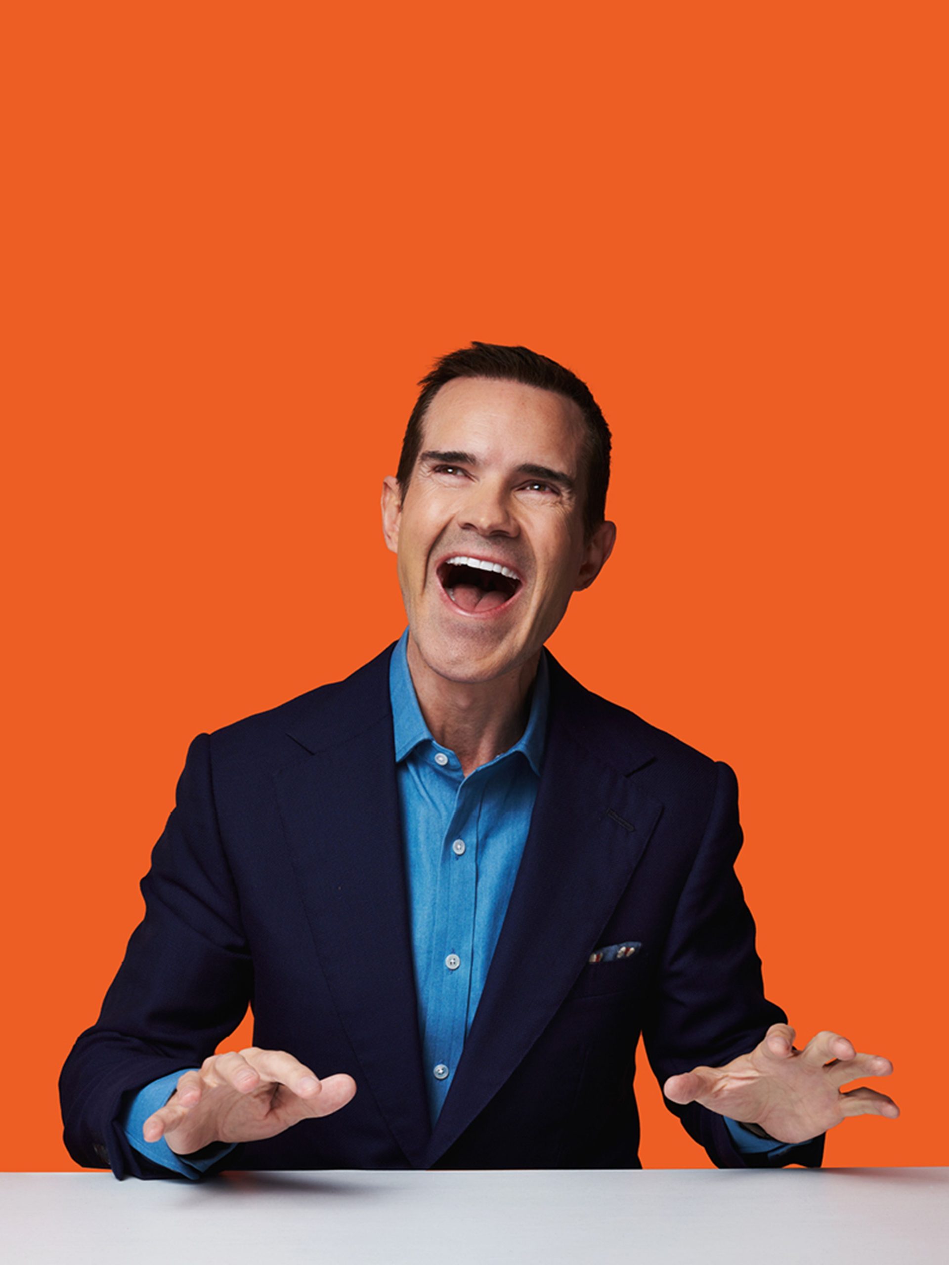 Man in a navy suit and blue shirt laughing with hands outstretched over a white table against an orange background.