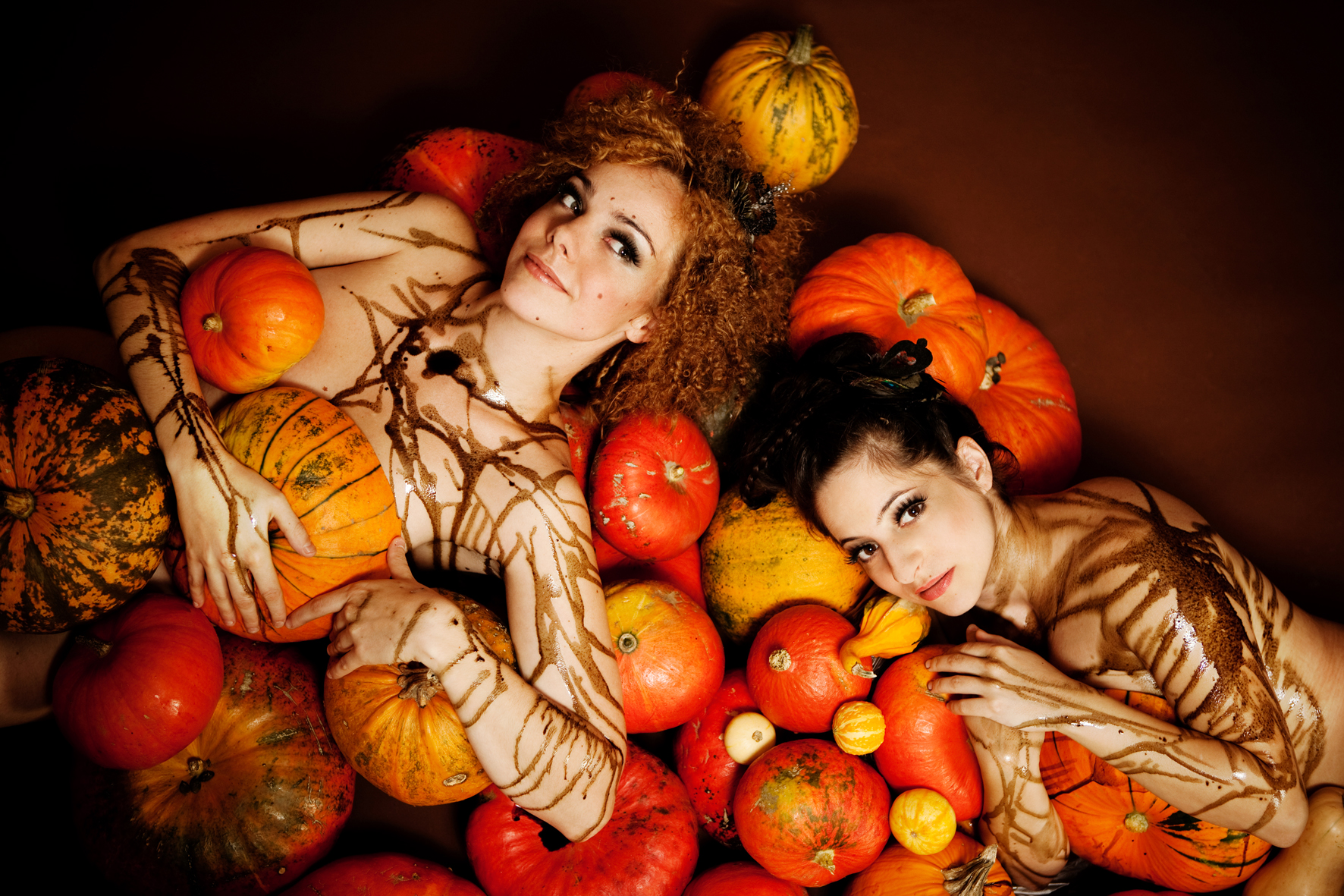 Two women lying among pumpkins, covered in brown vine-like body paint for a Halloween/ autumn theme.