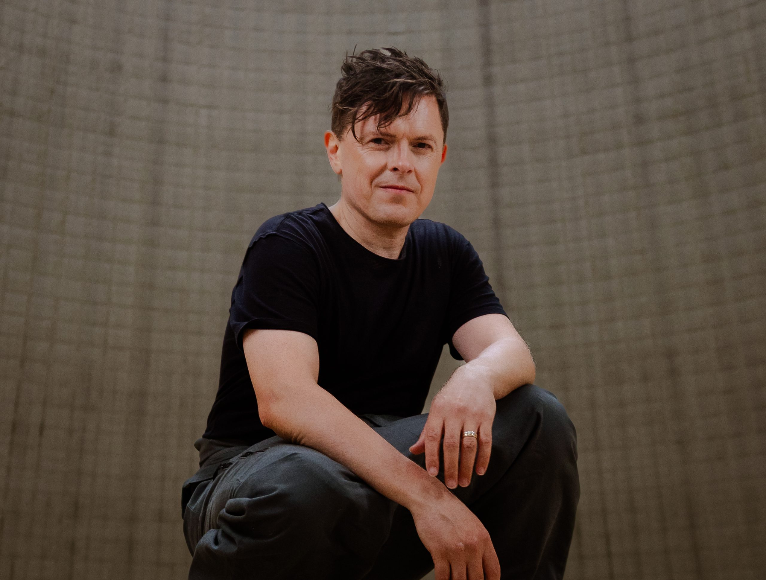 Man wearing a black T-shirt seated with arms resting on his knee against a beige textured wall.