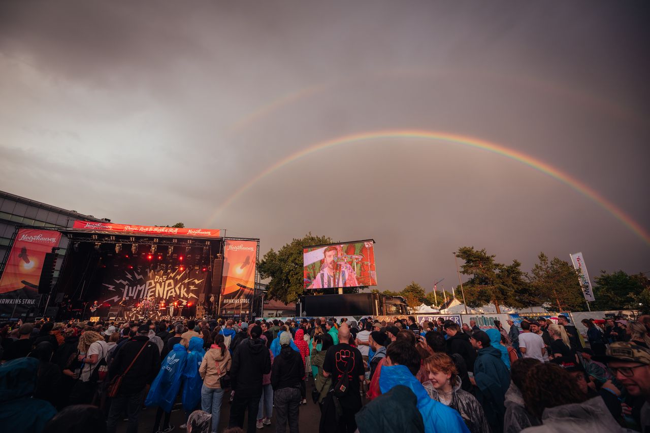 Crowd at an outdoor concert with a double rainbow arcing across a dark, cloudy sky behind the stage.