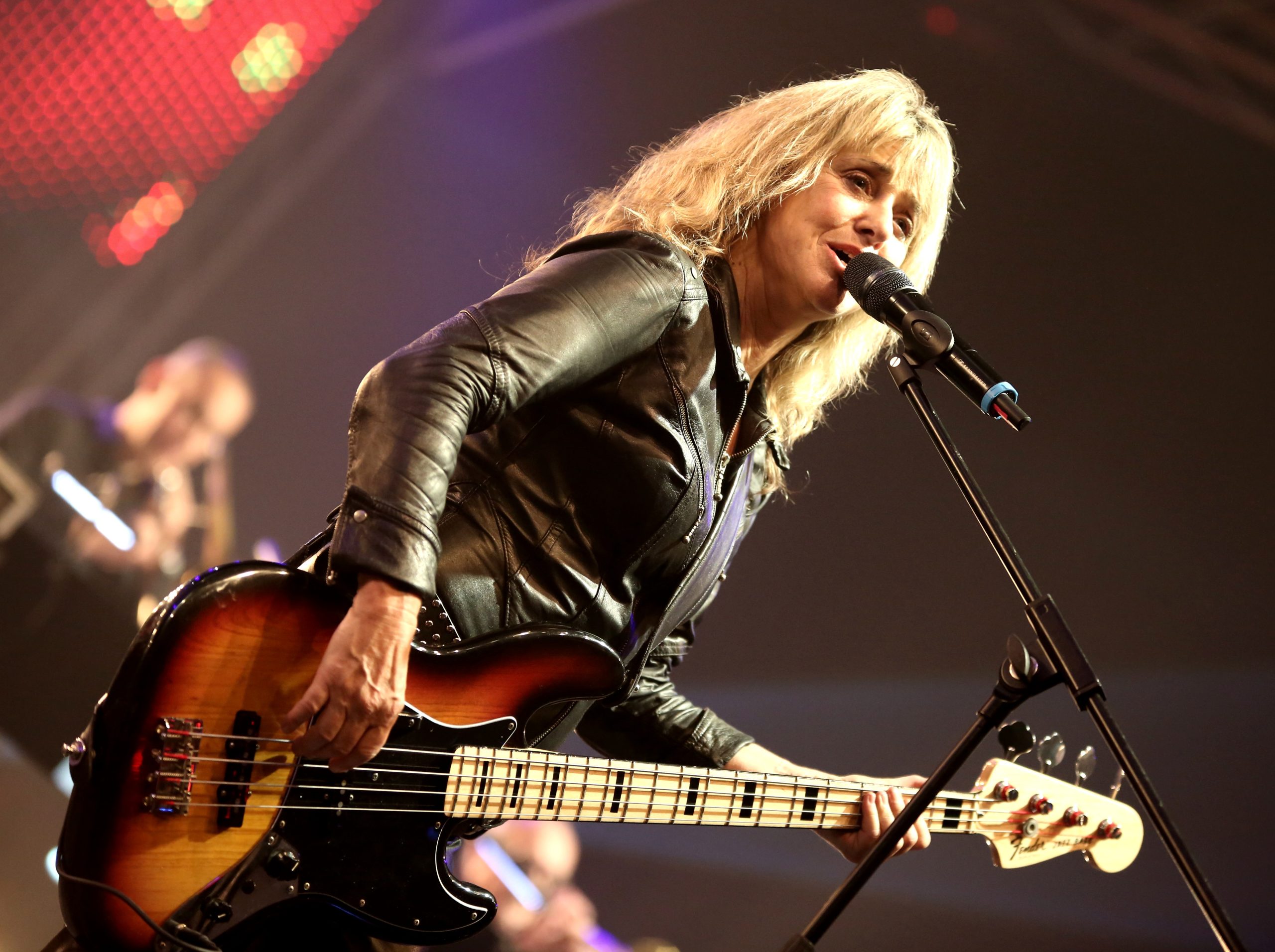 Female musician on stage playing a sunburst bass guitar, wearing a black leather jacket, singing into a microphone.